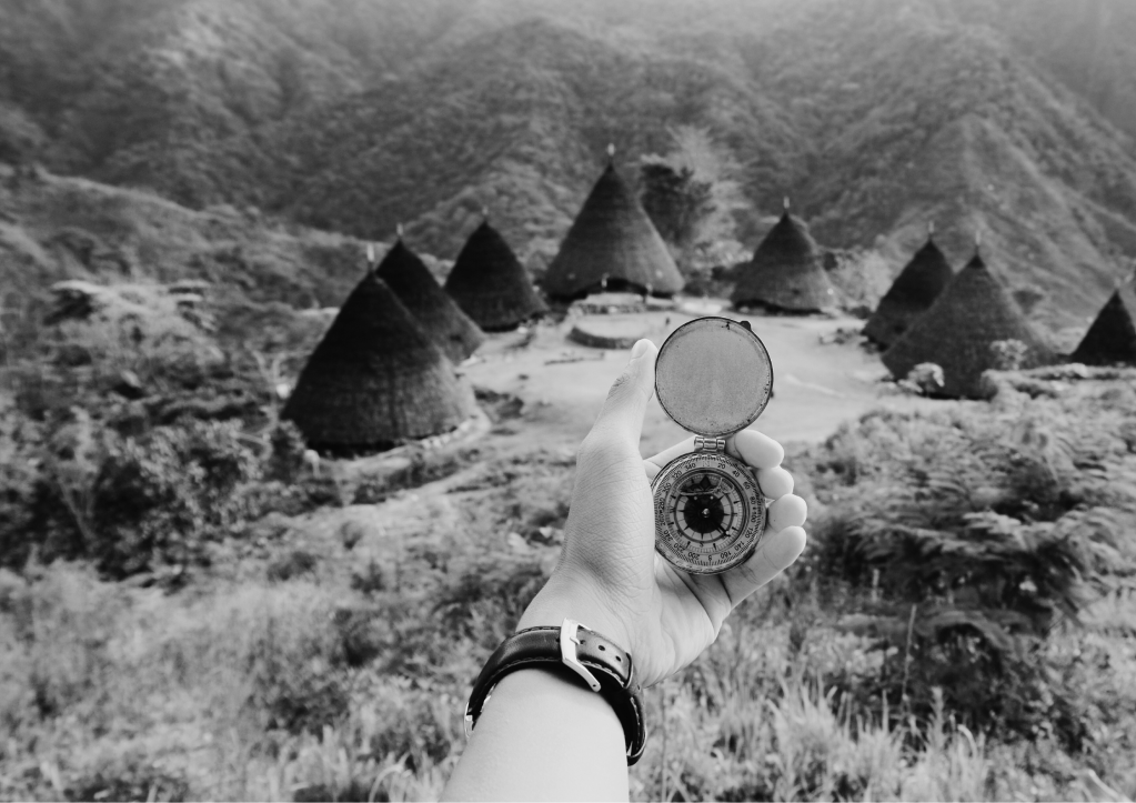 Main tenant une boussole devant des maisons traditionnelles dans un paysage montagneux, symbolisant l’exploration et la guidance